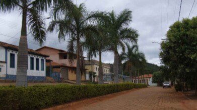 Processos Seletivos 7 Rua arborizada com palmeiras e casas em estilo simples na cidade de Olhos D’Água MG, com céu nublado e veículos estacionados.