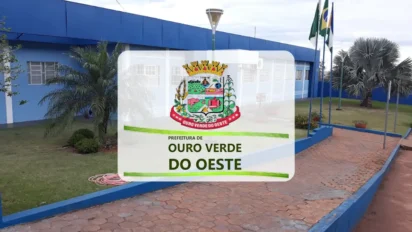 Fachada da Prefeitura de Ouro Verde do Oeste com prédio azul, bandeiras hasteadas, palmeiras e logomarca oficial centralizada sobre a imagem.