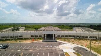 Vista frontal do prédio principal do PAX RN, com arquitetura moderna, estacionamento amplo e área verde ao fundo sob céu parcialmente nublado.