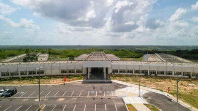 Processos Seletivos 9 Vista frontal do prédio principal do PAX RN, com arquitetura moderna, estacionamento amplo e área verde ao fundo sob céu parcialmente nublado.