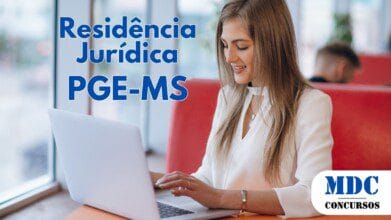 Processos Seletivos 4 Mulher jovem com cabelo castanho comprido, vestindo blazer branco, sorrindo enquanto digita em um laptop prateado em um café com assentos vermelhos e janelas grandes - PGE-MS abre inscrições para Residência Jurídica
