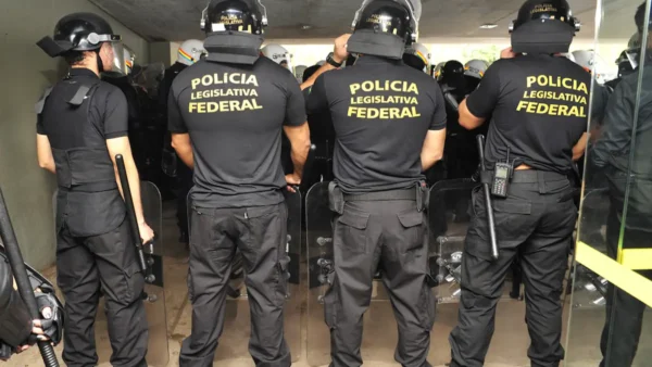 Agentes da Polícia Legislativa Federal vistos de costas, usando uniformes pretos, capacetes e escudos de proteção, posicionados em formação dentro de um prédio. Foto: Divulgação/Sindilegis
