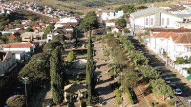 Processos Seletivos 8 Imagem aérea de uma praça arborizada no centro de Paraguaçu, Minas Gerais, com calçamento em padrão geométrico, bancos e postes de iluminação distribuídos ao longo da via central. No canto inferior esquerdo, há um coreto coberto com telhado de telhas claras. A praça é cercada por ruas com carros estacionados e ladeada por casas, prédios comerciais e uma igreja de grande porte ao fundo. Ao longe, é possível ver morros verdes e construções residenciais.