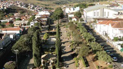 Imagem aérea de uma praça arborizada no centro de Paraguaçu, Minas Gerais, com calçamento em padrão geométrico, bancos e postes de iluminação distribuídos ao longo da via central. No canto inferior esquerdo, há um coreto coberto com telhado de telhas claras. A praça é cercada por ruas com carros estacionados e ladeada por casas, prédios comerciais e uma igreja de grande porte ao fundo. Ao longe, é possível ver morros verdes e construções residenciais.