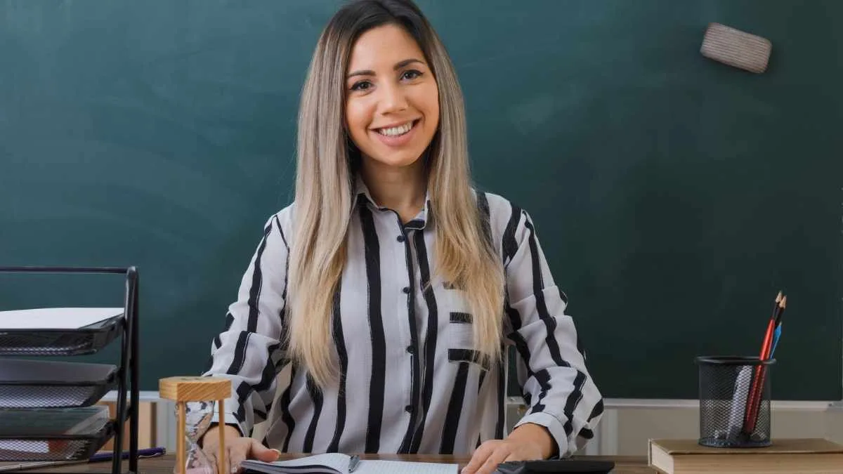 Imagem retrata uma mulher sorridente em sala de aula, com camisa listrada, sentada à mesa com materiais escolares, diante de um quadro verde, transmitindo profissionalismo e acolhimento. Foto: Freepik