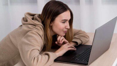 Processos Seletivos 7 Mulher jovem usando moletom bege olhando atentamente para a tela de um notebook em uma mesa.