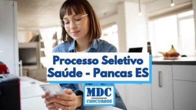 Imagem de uma jovem mulher de cabelos castanhos lisos e franja, usando óculos de grau e camisa jeans, sentada em uma cozinha moderna e iluminada, segurando um celular e sorrindo levemente enquanto olha para a tela, em primeiro plano há um texto em destaque escrito "Processo Seletivo Saúde - Pancas ES" em letras azuis com fundo branco e o logotipo "MDC Concursos" abaixo
