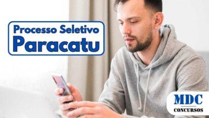 Homem jovem de barba curta e cabelo curto, vestindo moletom cinza com capuz, segura um smartphone enquanto olha para a tela. Ele está sentado em um ambiente iluminado, com um notebook branco à sua frente e uma cortina bege ao fundo - Paracatu Oferece 51 Vagas e Salário de R$ 2,8 mil para ACS em Processo Seletivo