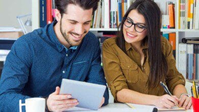 Homem e mulher estudando juntos em ambiente com livros, ele segura um tablet enquanto ela faz anotações sorrindo. Foto: nensuria/Freepik