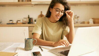 Mulher sorridente usando óculos estuda em frente ao notebook, com livros e caneca sobre a mesa.
