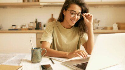 Mulher sorridente usando óculos estuda em frente ao notebook, com livros e caneca sobre a mesa.