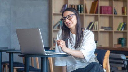 A imagem mostra uma jovem mulher de cabelos longos e lisos, usando óculos e vestindo uma camiseta branca, sentada em uma cadeira em frente a um notebook. Ela sorri enquanto segura uma caneta na mão direita e interage com a tela, sugerindo que está participando de uma videochamada ou estudando online. Ao fundo, há uma estante com livros organizados e o ambiente remete a uma biblioteca ou espaço de estudo moderno.