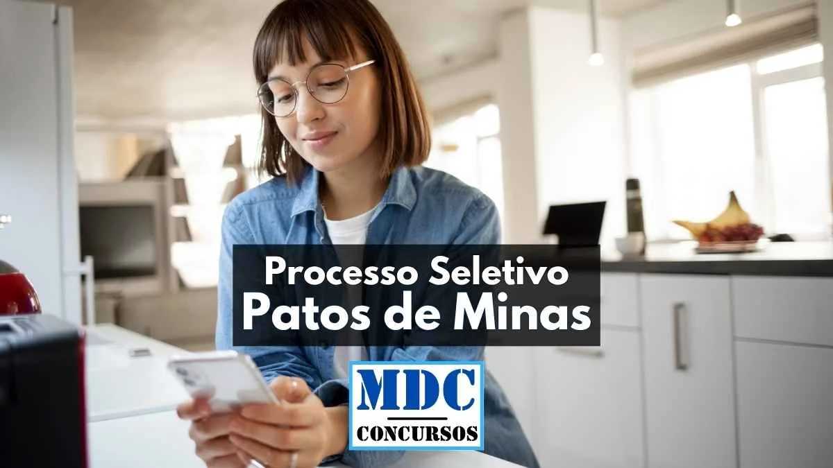 Mulher jovem usando óculos e blusa jeans manuseia um celular enquanto está sentada em uma cozinha moderna e iluminada. Na imagem, há sobreposição de texto com os dizeres “Processo Seletivo Patos de Minas” e o logotipo “MDC Concursos”. A cena transmite um momento de concentração, sugerindo que a candidata está realizando inscrição para o processo seletivo por meio de aplicativo.