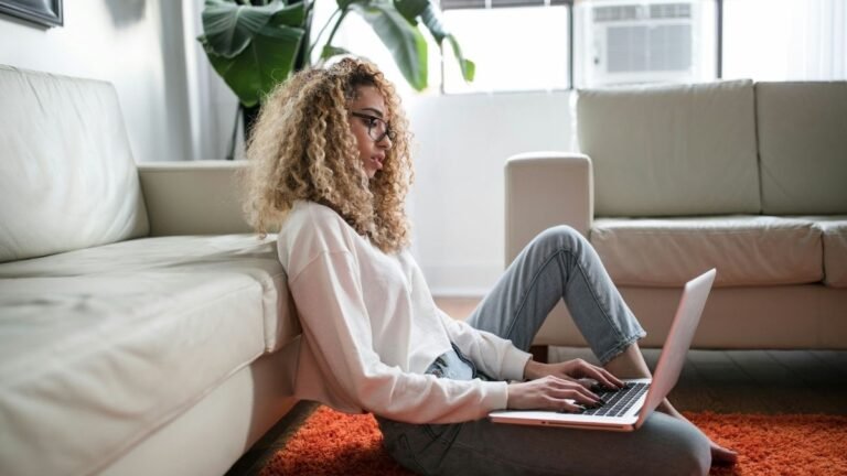 Mulher sentada no chão encostada no sofá usando notebook em ambiente iluminado com plantas ao fundo. Foto: Thought Catalog/Unsplash