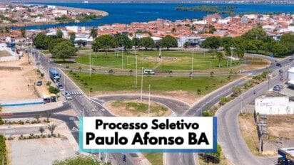 Vista aérea de uma grande rotatória arborizada em Paulo Afonso, na Bahia, com ruas asfaltadas ao redor, veículos em circulação e áreas residenciais ao fundo. A imagem destaca o urbanismo organizado da cidade, próxima a um lago visível no horizonte. Em primeiro plano, há uma faixa informativa com os dizeres “Processo Seletivo Paulo Afonso BA”, indicando chamada para seleção pública no município.