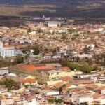 Vista aérea de uma cidade com prédios baixos, casas, uma igreja azul ao centro-esquerda e uma chaminé alta de tijolos à direita. Ao fundo, áreas rurais e colinas.
