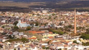 Vista aérea de uma cidade com prédios baixos, casas, uma igreja azul ao centro-esquerda e uma chaminé alta de tijolos à direita. Ao fundo, áreas rurais e colinas.
