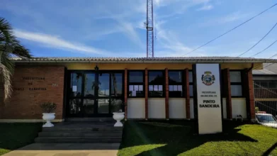 Fachada da Prefeitura Municipal de Pinto Bandeira, um prédio térreo de tijolos à vista e amplas janelas de vidro, com um totem branco à direita exibindo o brasão e o nome da instituição sob um céu azul. Foto: Divulgação/Google Maps (Teresinha Romano)