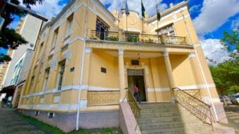 Fachada do prédio da Prefeitura Municipal de Poços de Caldas, em estilo histórico, com escadaria, colunas e bandeiras hasteadas.