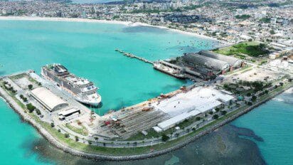 Vista aérea do Porto de Maceió com navio de cruzeiro atracado, galpões industriais, mar azul e parte urbana da cidade ao fundo.