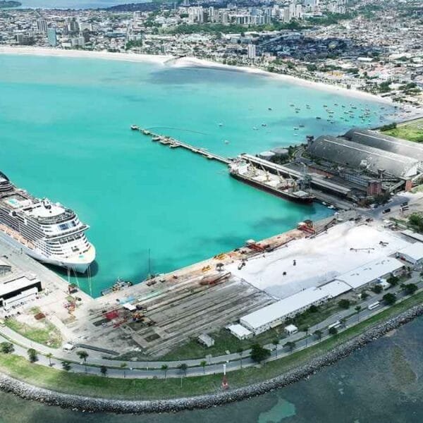 Vista aérea do Porto de Maceió com navio de cruzeiro atracado, galpões industriais, mar azul e parte urbana da cidade ao fundo.