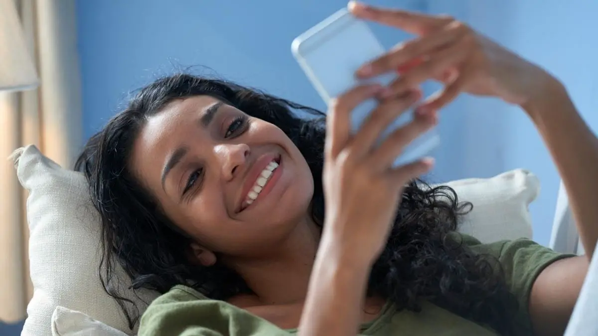 Mulher sorridente deitada em um sofá usando um celular. Foto: pressfoto/Freepik