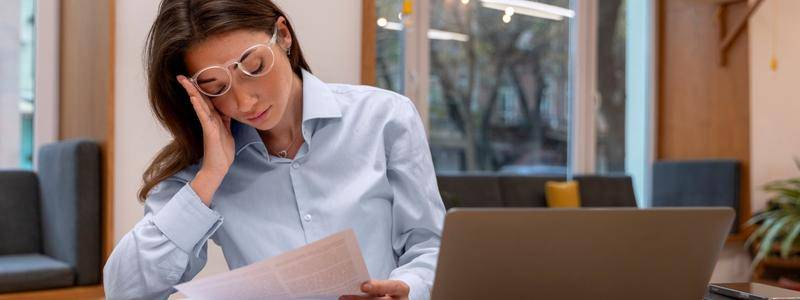 Como estudar legislação seca com eficiência sem precisar decorar tudo 2 Mulher de óculos e camisa azul, com uma mão na cabeça, estuda concentrada segurando um documento e olhando para um laptop. Foto: Freepik