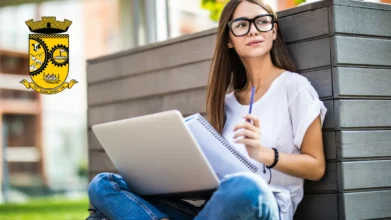 Mulher sentada ao ar livre usando notebook e caderno, em ambiente urbano, com o brasão do município de Agrolândia ao lado, representando estudo e concurso público. Foto: diana.grytsku/Freepik