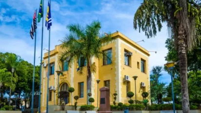 Prédio amarelo da Prefeitura de Agudos cercado por jardins, palmeiras e postes, com bandeiras hasteadas à frente sob céu azul. Foto: Divulgação/Prefeitura de Agudos-SP