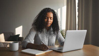 Processos Seletivos 6 Mulher sentada à mesa usando notebook e fazendo anotações em caderno, com uma caneca ao lado. Foto: karlyukav/Freepik