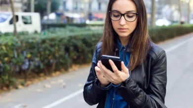 Mulher jovem usando óculos e jaqueta preta olha para o celular enquanto caminha por uma calçada arborizada em área urbana. Foto: nensuria/Freepik