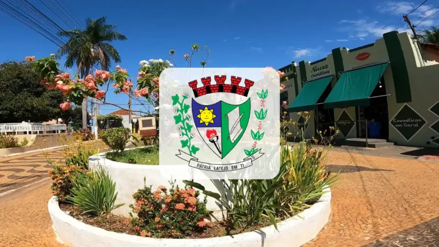 Fotografia de praça pública com canteiro circular de flores sob céu azul, destacando o brasão da cidade de Bilac ao centro e fachada comercial ao fundo