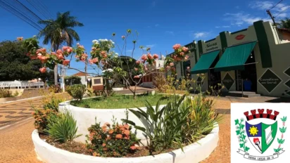 Canteiro central circular com flores em tons de salmão e folhagens verdes em uma praça urbana, com estabelecimentos comerciais ao fundo e o brasão de Bilac inserido no canto inferior direito. Foto: Divulgação/Prefeitura de Bilac-SP