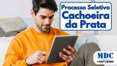 Homem jovem de barba curta e cabelo escuro, vestindo um moletom laranja, sentado no chão encostado na cama enquanto usa um tablet. O ambiente é um quarto com cama, almofadas e luminária moderna ao fundo - Prefeitura de Cachoeira da Prata (MG) oferece 14 vagas e salários de até R$ 5,3 mil em Processo Seletivo