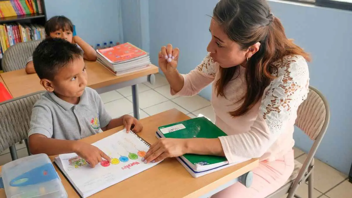 Professora auxilia criança com atividade escolar em sala de aula; ao fundo, outra criança observa sentada em sua carteira.