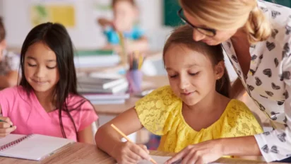 Professora auxiliando uma aluna em sala de aula enquanto outra criança escreve ao lado, em ambiente escolar com materiais sobre a mesa. Foto: gpointstudio/Freepik