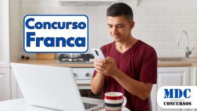 Homem de camiseta vermelha sentado em uma cozinha moderna, sorrindo enquanto usa um smartphone. Um laptop branco está aberto na mesa, ao lado de uma caneca listrada. Ao fundo, há um fogão, tábua de corte e torneira branca - Prefeitura de Franca oferece salários de até R$ 7 mil em 5 Concursos Públicos
