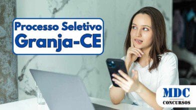 Mulher jovem de cabelos castanhos e pele clara sentada à mesa com um laptop aberto à sua frente, segurando um smartphone na mão direita e apoiando a esquerda no rosto, com expressão pensativa. O ambiente tem parede marmorizada ao fundo e iluminação natural - Prefeitura de Granja CE oferece salários de até R$ 3,5 mil em Processo Seletivo