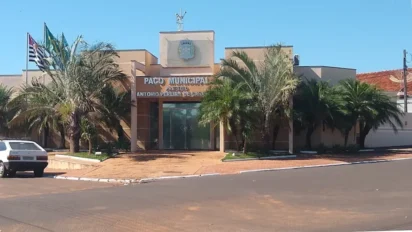 Fachada do Paço Municipal de Guzolândia, prédio de cor bege com palmeiras na frente, bandeiras hasteadas à esquerda e um carro branco estacionado. No centro, o letreiro identifica o edifício.