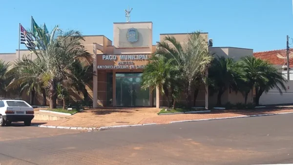 Fachada do Paço Municipal de Guzolândia, prédio de cor bege com palmeiras na frente, bandeiras hasteadas à esquerda e um carro branco estacionado. No centro, o letreiro identifica o edifício.