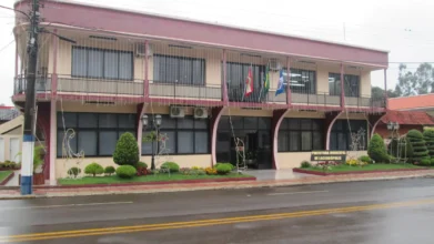 Fachada frontal da Prefeitura Municipal de Lacerdópolis em Santa Catarina, um prédio de dois pavimentos em tons de bege e bordô com bandeiras hasteadas na sacada e decorações natalinas no jardim frontal sob dia chuvoso.