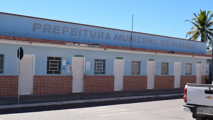 Fachada da Prefeitura Municipal de Mambaí, em Goiás, um prédio térreo azul claro com base de tijolos aparentes, diversas portas brancas e céu limpo ao fundo.