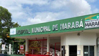 Fachada do Hospital Municipal de Marabá, no estado do Pará, com destaque para a placa verde com letras metálicas que identificam a instituição. À direita, há o logotipo da Prefeitura de Marabá com a frase "Um Governo por Todos". O céu está parcialmente nublado e parte da vegetação e portão de entrada do hospital também são visíveis na imagem.