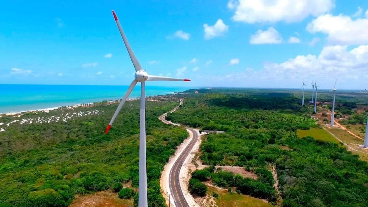 Aeroportas de energia eólica próximas ao litoral com estrada cercada por vegetação e mar ao fundo. Foto: Divulgação/Prefeitura de Mataraca-PB