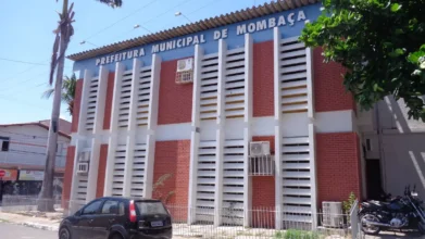 Fachada da Prefeitura Municipal de Mombaça com paredes de tijolos aparentes, grades brancas verticais, ar-condicionados e carros estacionados em frente. Foto: Divulgação/Prefeitura de Mombaça-CE