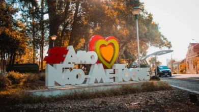 Monumento com a frase #Amo Não-Me-Toque em letras brancas e um coração estilizado em vermelho, amarelo e laranja, localizado em uma praça arborizada sob a luz do sol.