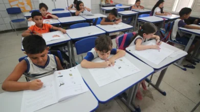 Crianças sentadas em carteiras escolares em sala de aula, concentradas realizando atividades em livros e cadernos. Foto: Divulgação/Prefeitura de Natal /RN