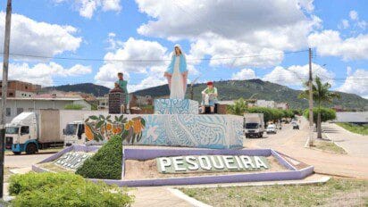 Imagem de uma rotatória na cidade de Pesqueira, Pernambuco, com destaque para um monumento central decorado com arte colorida e uma escultura da Virgem Maria ao centro, ladeada por figuras representando trabalhadores. O pedestal é adornado com pinturas de flores e frutas. Ao fundo, vê-se uma paisagem com morros verdes, céu azul com nuvens e uma avenida com caminhões e carros circulando.