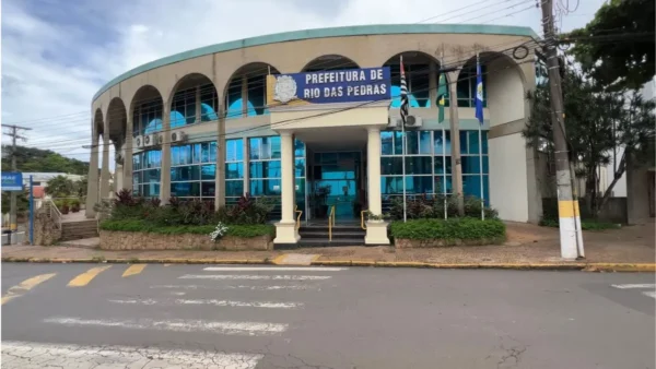 Prefeitura de Rio das Pedras/SP divulga resultado preliminar de concurso 2 Fachada da Prefeitura de Rio das Pedras com prédio de dois andares, arcos na parte superior, vidros azuis e bandeiras hasteadas na entrada. Foto: Divulgação/Prefeitura de Rio das Pedras-SP