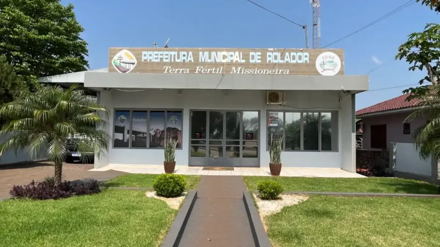 Fachada da Prefeitura Municipal de Rolador, no Rio Grande do Sul. O prédio térreo cinza possui gramado frontal, palmeiras laterais e um letreiro com a frase Terra Fértil Missioneira sob o céu azul.